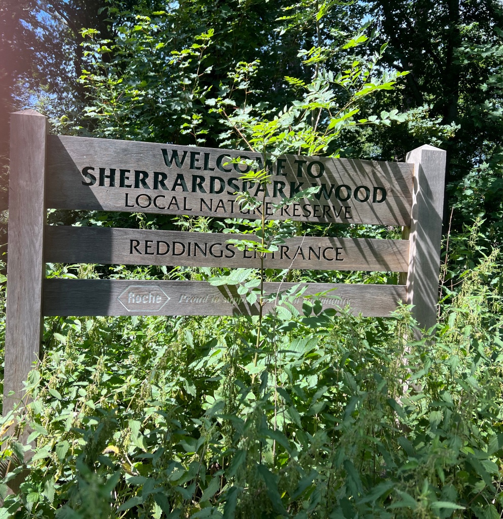 The wooden sign has 2 uprights with 3 planks horizontally across the uprights. Each plank has text carved upon it.

The top plank is the widest and has 3 lines of text that reads “Welcome To
Sherrardspark Wood
Local Nature Reserve”
The middle plank reads “Reddings Entrance”
The bottom plank reads “Roche Proud to support our local community”
The green vegetation is partially obscuring the text on each plank.